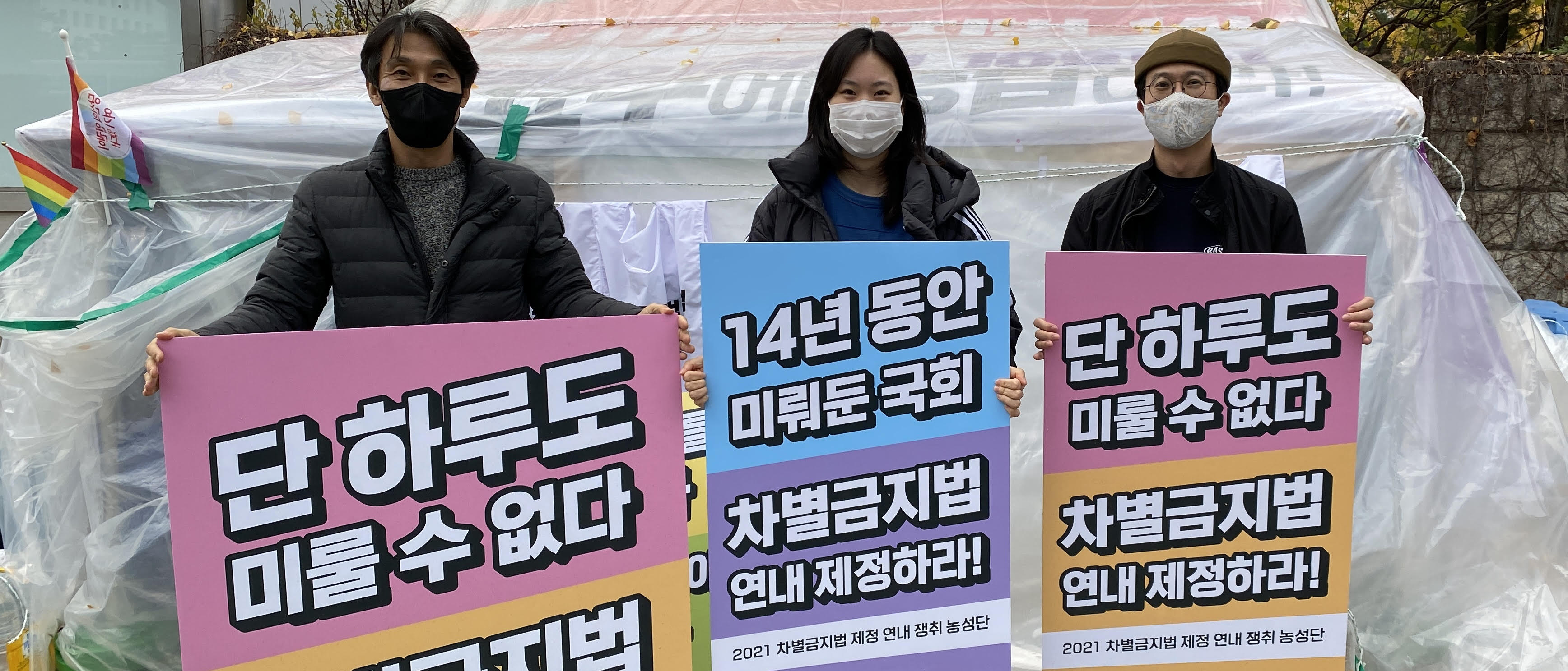 “국회 앞에 설치된 차별금지법 연내 제정 농성장 앞에서 세 명의 사람이 피켓을 들고 서 있어요. 피켓에는 단 하루도 미룰 수 없다, 14년 동안 미뤄둔 국회 차별금지법 연내 제정하라 라는 글이 쓰여 있습니다.“